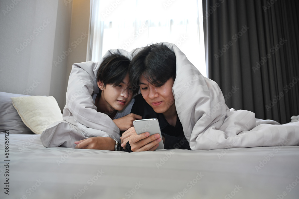 Young gay couple lying under blanket on bed and using smart phone together. LGBT and love concept