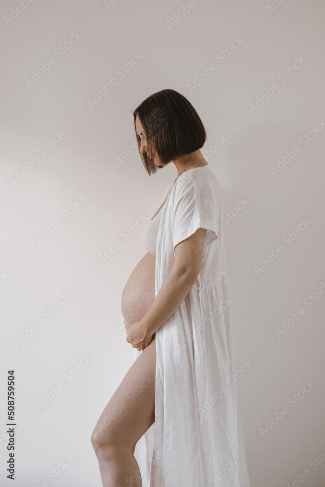 Maternity happiness concept. Young pretty happy pregnant woman in elegant white robe dress touch tummy. Minimalist aesthetic pregnancy