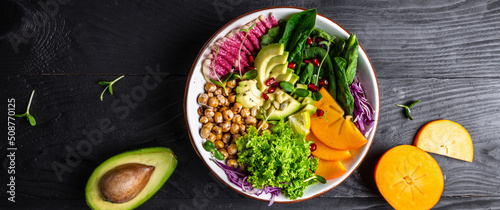 Fotografering Fresh salad with roasted chickpeas, avocado, persimmon, spinach, avocado, watermelon radish and seeds on a dark background