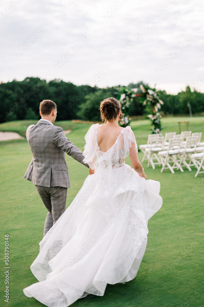 Bride and groom walking and holding hands on a green meadow. bridal dress in boho style