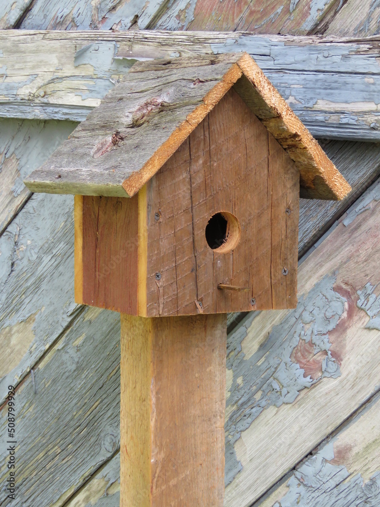 Simple Natural Wooden Birdhouse on Wood Post in Front of Old Weathered Gray Barn with Peeling Grey Paint