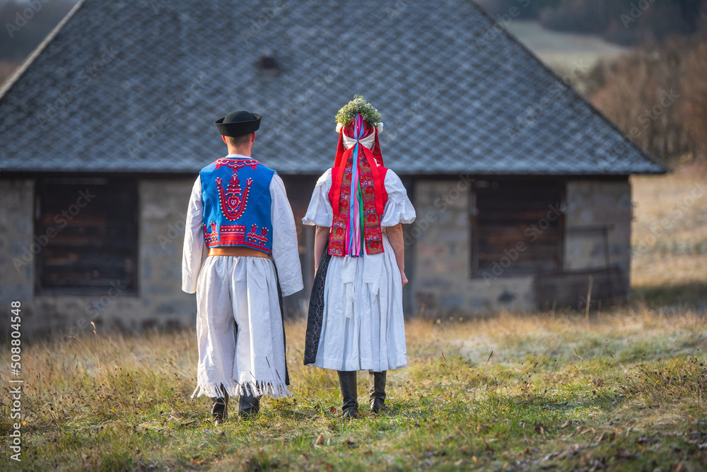 A couple dressed in traditional folk costume. Slovak costume in autumn nature. Old country cottage in the background. Young couple in folk costume walking in the garden A couple dressed in traditional folk costume. Slovak costume in autumn nature. Old country cottage in the background. Young couple in folk costume walking in the garden