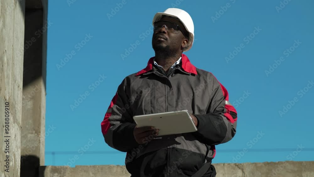 African american worker stands at construction site and inputs data in his tablet computer