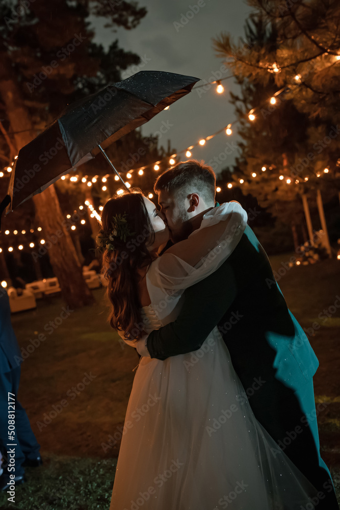 A couple in love under a black umbrella kisses. Curvy people, wedding couple on a walk. Stylish charming bride, groom in green suit. Beautiful park A couple in love under a black umbrella kisses. Curvy people, wedding couple on a walk. Stylish charming bride, groom in green suit. Beautiful park