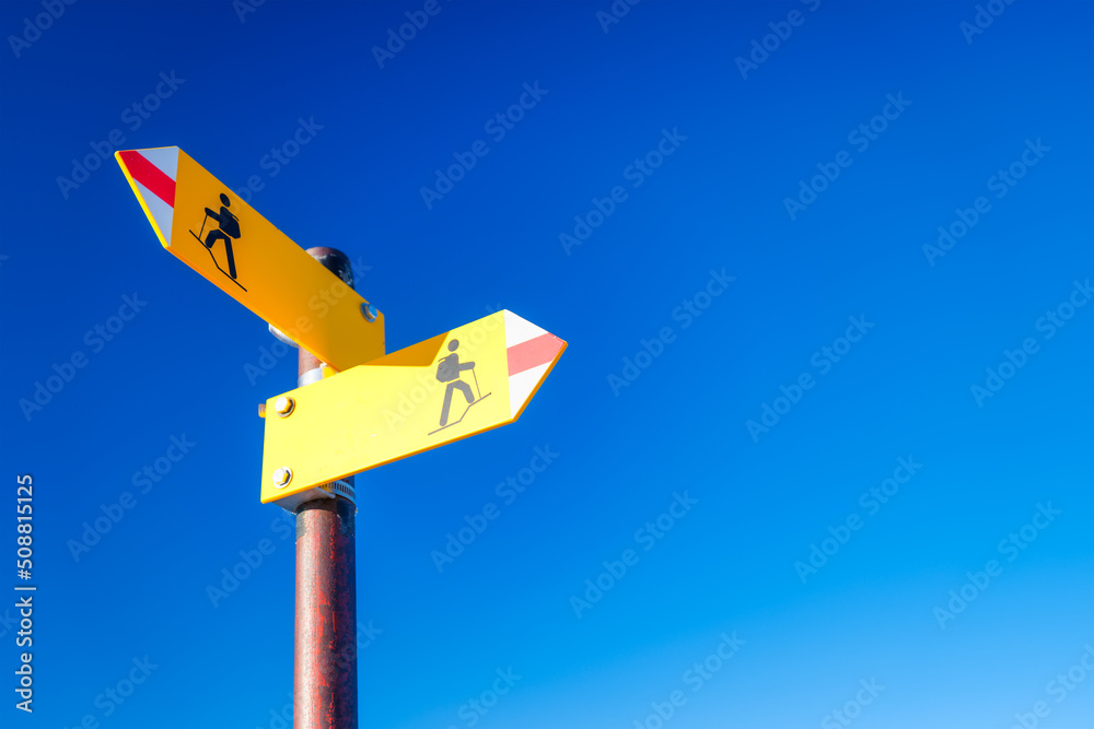 Directional marker for a mountain hike. Yellow bright sign with an arrow against the blue sky. Navigation in a hiking trip. Directional marker for a mountain hike. Yellow bright sign with an arrow against the blue sky. Navigation in a hiking trip.