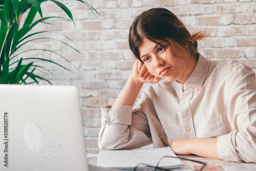 Young woman student sitting at desk full with books textbooks looking at computer screen to unfinished homework feels tired bored and unmotivated, girl loaded with tasks, work backlog overflow concept photo