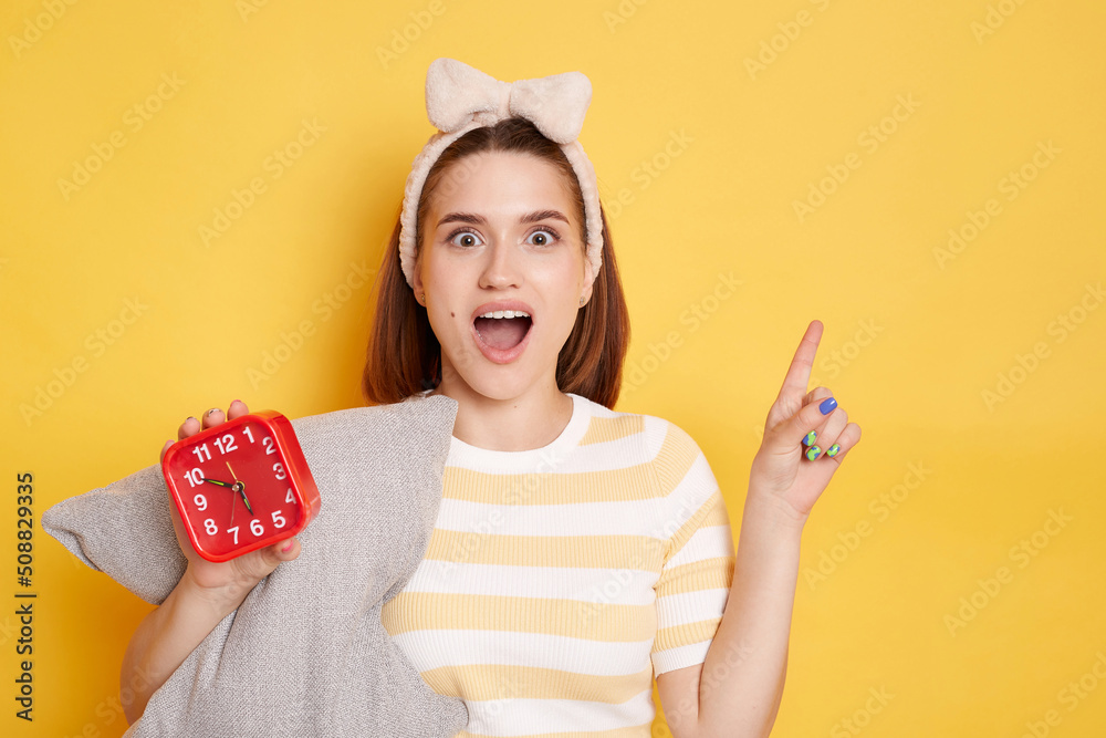 Portrait of excited woman wearing t shirt and hair band posing isolated on yellow background, holding pillow and red alarm clock, looking at camera with excitement, pointing aside at copy space. Portrait of excited woman wearing t shirt and hair band posing isolated on yellow background, holding pillow and red alarm clock, looking at camera with excitement, pointing aside at copy space.