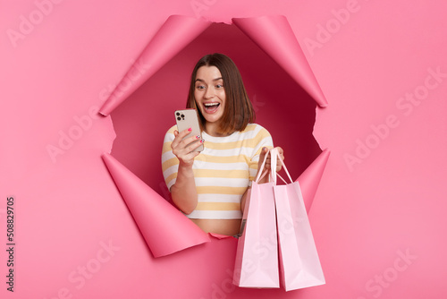 Image of excited surprised woman wearing striped shirt posing in breakthrough of pink background, standing with shopping bags and smart phone, checking cashback. photo