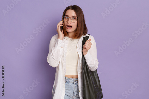 Portrait of shocked beautiful woman with black bag wearing white shirt and jeans posing isolated over purple background, talking phone, hearing shocking news, looking at camera with open mouth,