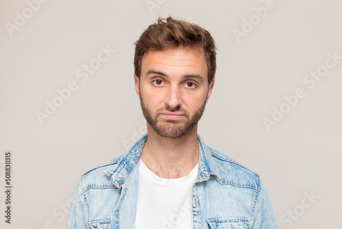 Young hipster male with stubble in a white t-shirt and light blue denim shirt. Emotion - a dissatisfied face, disgust, sees something unpleasant. On a light gray background
