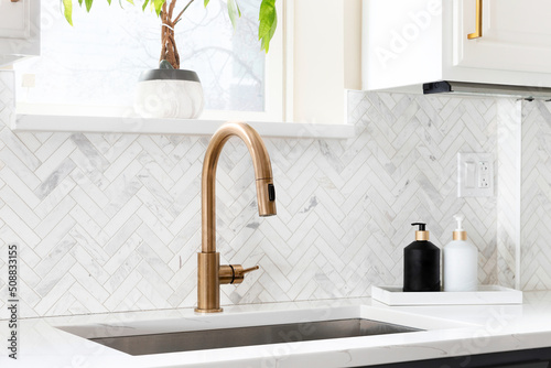 Sink detail shot in a luxury kitchen with herringbone backsplash tiles. white marble countertop, and gold faucet.	