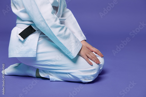 Cropped view of child aikido wrestler in white kimono sitting in stance with hands on knees isolated on purple background with copy space. Concept of Oriental Martial Arts practice