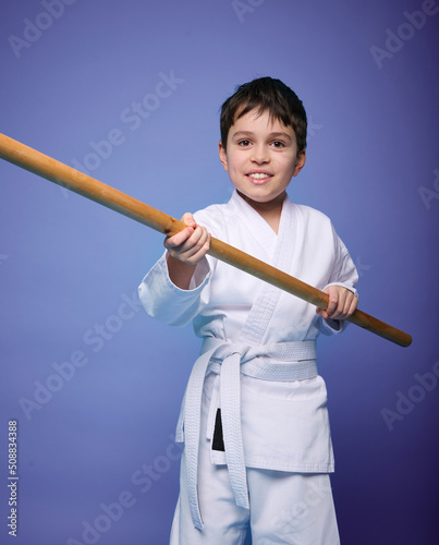 Confident strong concentrated boy - Aikido wrestler in white kimono stands with a wooden jo weapon in his hands. Oriental martial arts practice concept