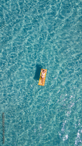 Aerial vertical view of woman relaxing and enjoying summer holiday vacation laying on a colorful inflatable lilo mattress on blue water surface. People enjoying ocean travel destination. Vertical