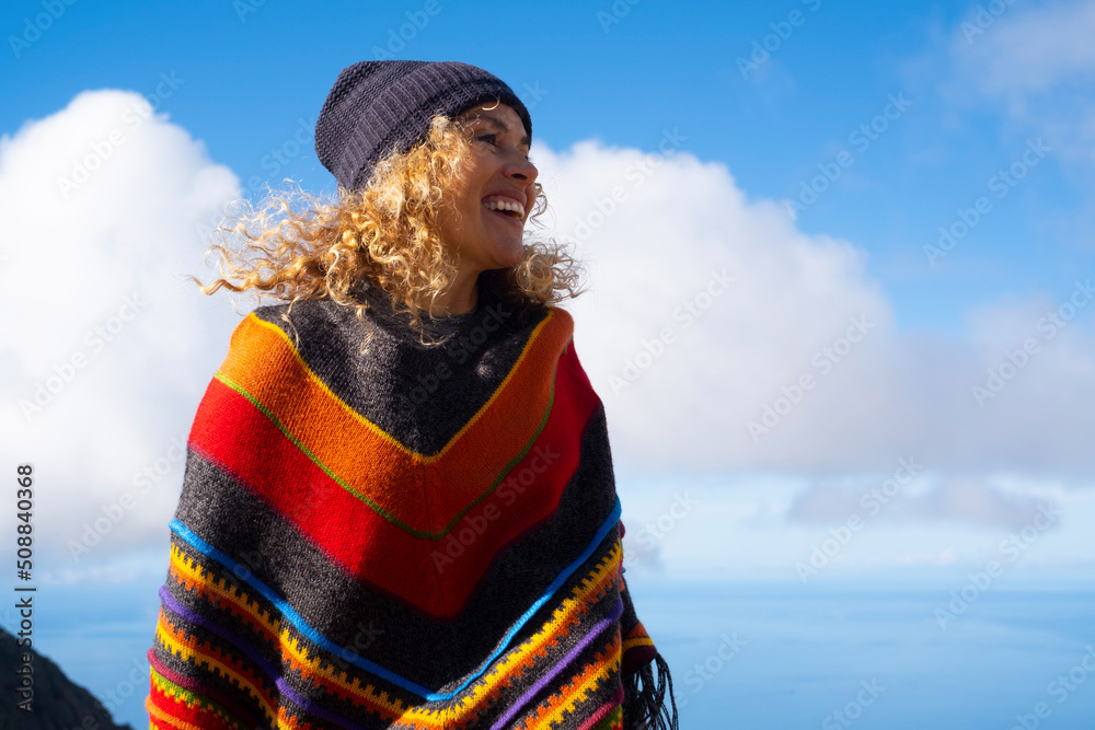Colorful poncho clothes on happy pretty adult woman enjoying outdoor leisure activity against a blue sky background. Happy and cheerful people female portrait with winter hat - obrazy, fototapety, plakaty 
