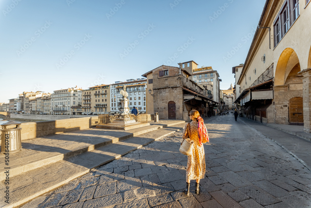 Fototapeta premium Woman walking on famous old bridge, called Ponte Vecchio, in Florence. Concept of traveling italian landmarks. Stylish woman with colorful shawl and sunglasses. Wide view on sunrise