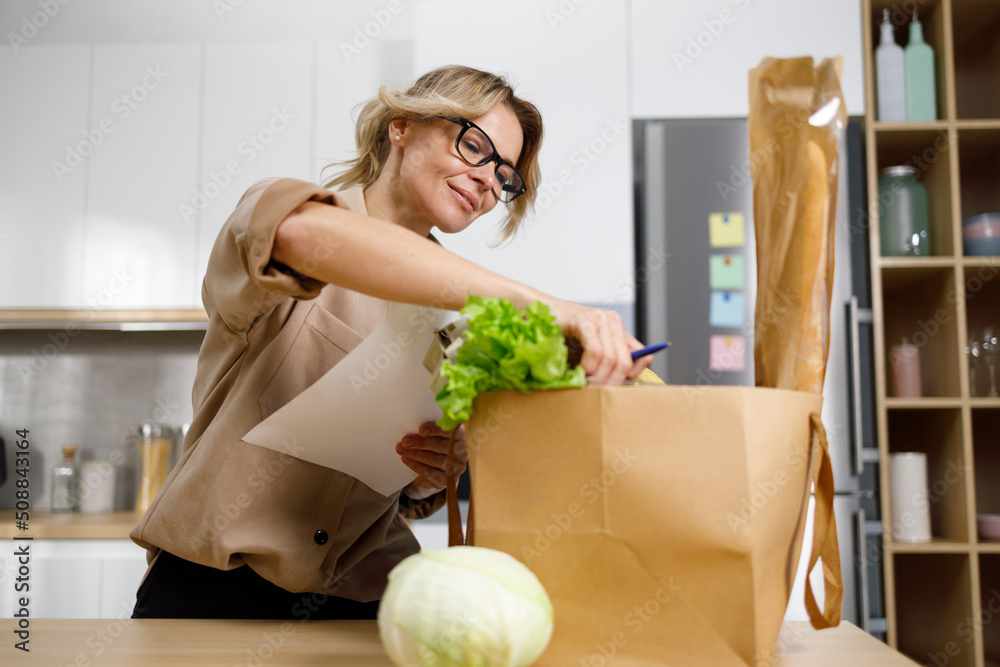 Middle-aged housewife checks purchased groceries against a list. Woman keeps track of food costs Middle-aged housewife checks purchased groceries against a list. Woman keeps track of food costs