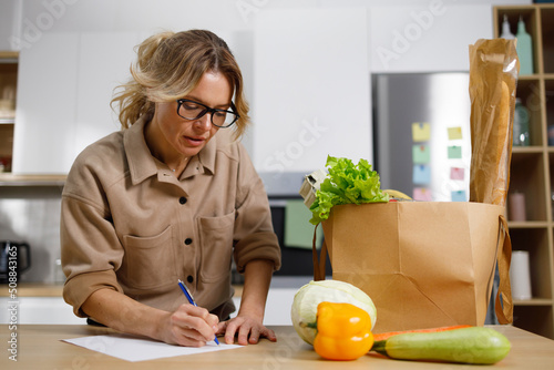 Pretty mature woman is checking shopping list at the table in the kitchen. Food cost control concept