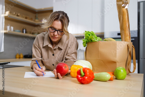 Mature housewife writes down expenses for purchased food. Woman in the kitchen and a paper bag with grocery