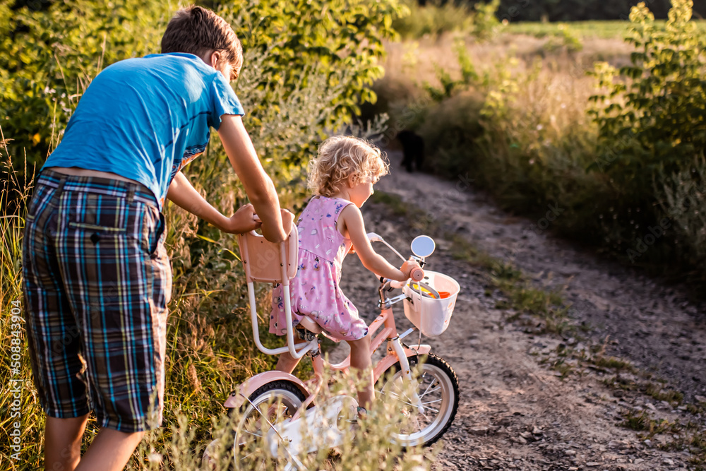 brother teaches little sister to ride bicycle. girl rides along rural road on two-wheeled bicycle among summer grasses