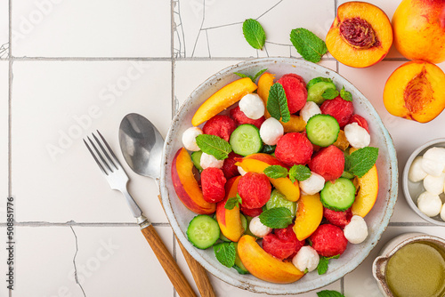 Summer fruit salad with mozzarella cheese, cucumber and mint in a plate with cutlery on white tile table. top view