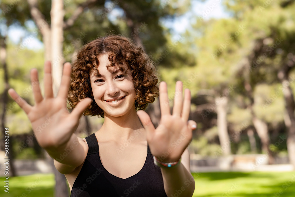 Happy redhead woman wearing black sports bra standing on city park, outdoor showing and pointing up with fingers number nine while smiling confident and happy. Selective focus on her face. Happy redhead woman wearing black sports bra standing on city park, outdoor showing and pointing up with fingers number nine while smiling confident and happy. Selective focus on her face.
