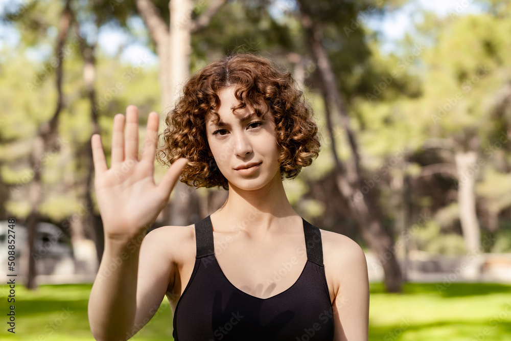 Portrait of young redhead woman wearing sports bra standing on city park, outdoor doing stop sing with palm of the hand. Warning expression with negative and serious gesture on the face.