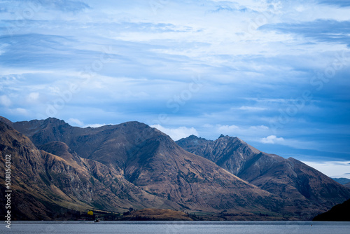 Jagged form of Remarkables Mountain Range across Lake Wakatipu without snow and shadows and light through overcast sky