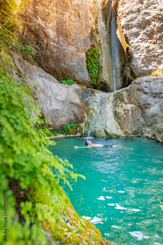 Young happy man floating in turquoise blue crystal clear water in the river with a waterfall summer vacation relaxing body and mind.