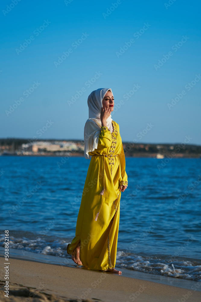 Arab woman with hijab and yellow Moroccan dress on the beach, enjoying her summer vacation. Walking along the shore. Vertical photo Arab woman with hijab and yellow Moroccan dress on the beach, enjoying her summer vacation. Walking along the shore. Vertical photo