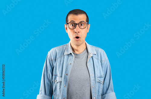 Studio shot of a hispanic man wearing glasses over isolated blue background afraid and shocked with surprise expression, fear and excited face.