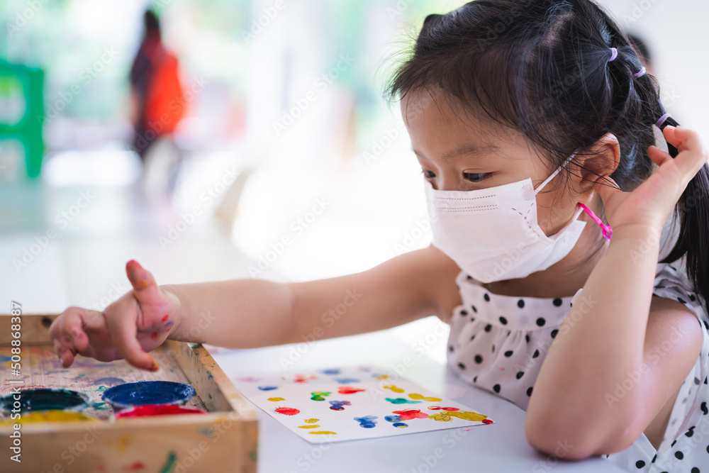 Portrait image child 5 years old. Little girl wearing white medical face mask to prevent toxic dust pm2.5 and virus. Cute kid doing art craft with her finger. Learning water color with my body concept Portrait image child 5 years old. Little girl wearing white medical face mask to prevent toxic dust pm2.5 and virus. Cute kid doing art craft with her finger. Learning water color with my body concept
