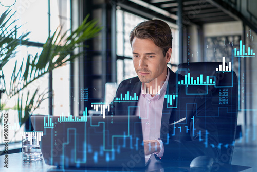 Handsome businessman in suit at workplace working with laptop to optimize trading strategy at corporate finance fund. Forex chart hologram over office background photo