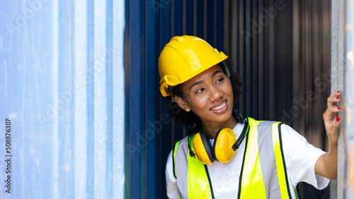 Young African American trainee in safety suit, yellow hard hat and eye muff stand smiling next to big container in a shipyard. African curly hair female worker in smile. Female loader with eye muff