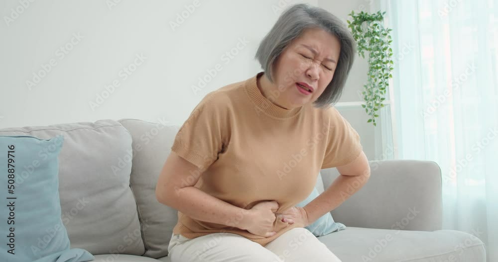 Beautiful elderly Asian woman sits on the sofa, hand clutching her stomach due to severe abdominal pain,diarrhea, gastritis, enteritis in the living room alone.People's health problems concept. Beautiful elderly Asian woman sits on the sofa, hand clutching her stomach due to severe abdominal pain,diarrhea, gastritis, enteritis in the living room alone.People's health problems concept.