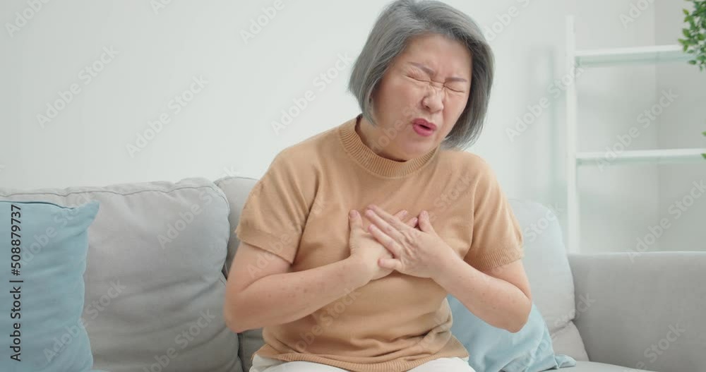 Asian Senior old Asian woman suffer from heart attack sitting on sofa in living room at home.Elder female with suffering and chest pain has difficulty breath clutching her chest from acute pain. Asian Senior old Asian woman suffer from heart attack sitting on sofa in living room at home.Elder female with suffering and chest pain has difficulty breath clutching her chest from acute pain.
