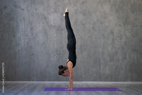 Side view portrait beautiful young woman wearing black sportswear, exercising against a gray wall doing yoga or pilates. Standing in a pose. Full length