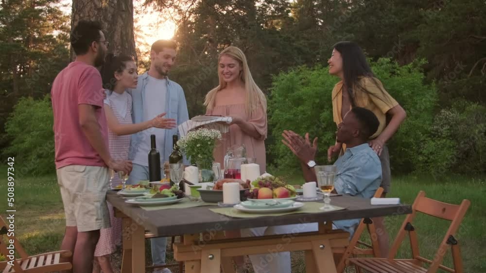 Group of multi-ethnic young friends spending time together in local park having party, young woman bringing pie to table Group of multi-ethnic young friends spending time together in local park having party, young woman bringing pie to table