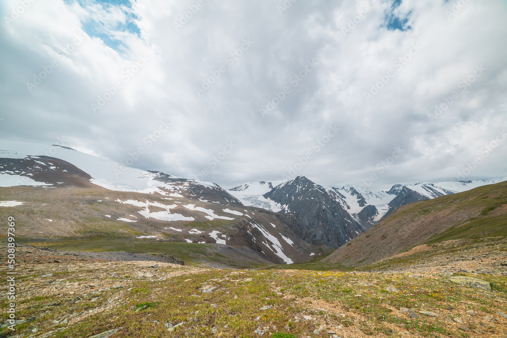 Scenic panoramic view from sunlit green grassy hill to high snowy mountain range with sharp tops and glaciers under gray cloudy sky. Colorful landscape with large snow mountains at changeable weather.