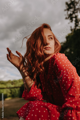 Portait of beauty young redhead woman with long curly thick hair dressed in red shirt dress with white dots posing on a camera in sunlight