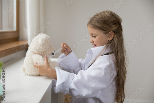 Cute little girl in white coat play pediatrician, pretend be doctor treats best fluffy friend, holds syringe makes vaccine or injection to stuffed toy bear. Healthcare, medicine, vet clinic ad concept