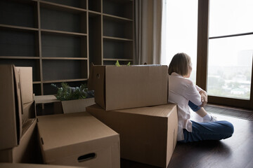 Rear back view young woman sit on floor near heap of cardboard boxes on relocation day. Move-out to rented house, divorced female start new life alone look out window feels depressed. Eviction concept