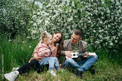  family mom mom baby daughter in the garden blooming apple trees, father playing the ukulele