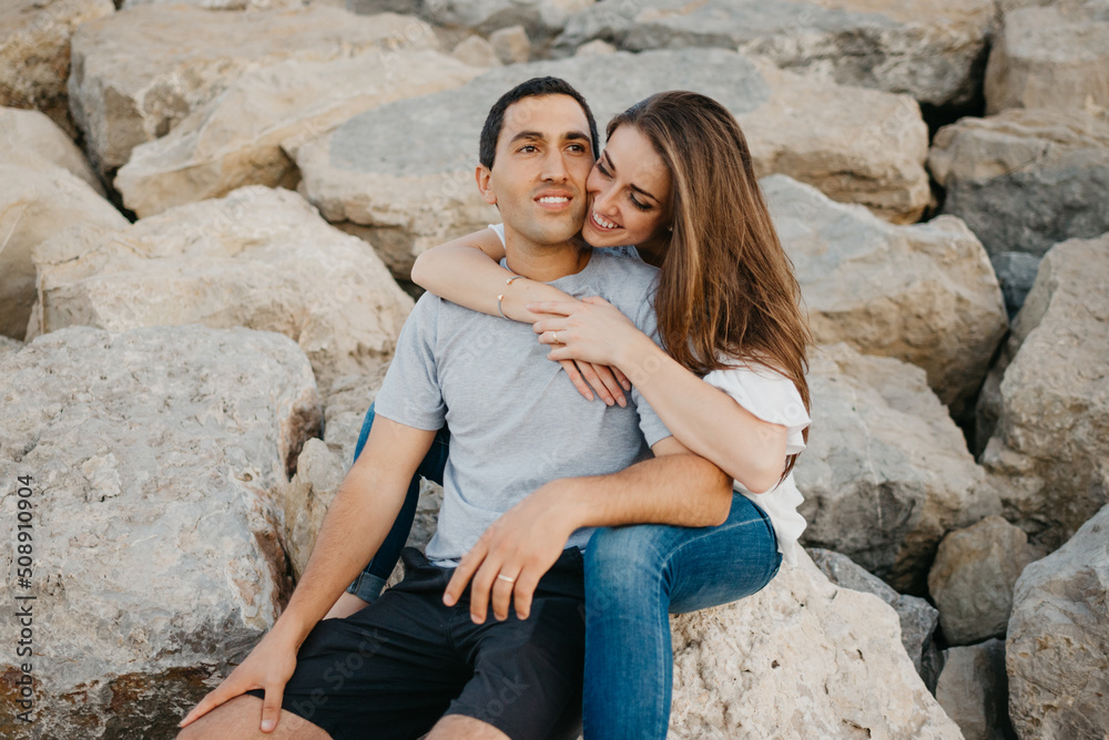 A Hispanic man is sitting between the legs of his smiling girlfriend on the stones of a breakwater in Spain in the evening. A happy woman is hugging her husband on a date at sunset in Valencia.