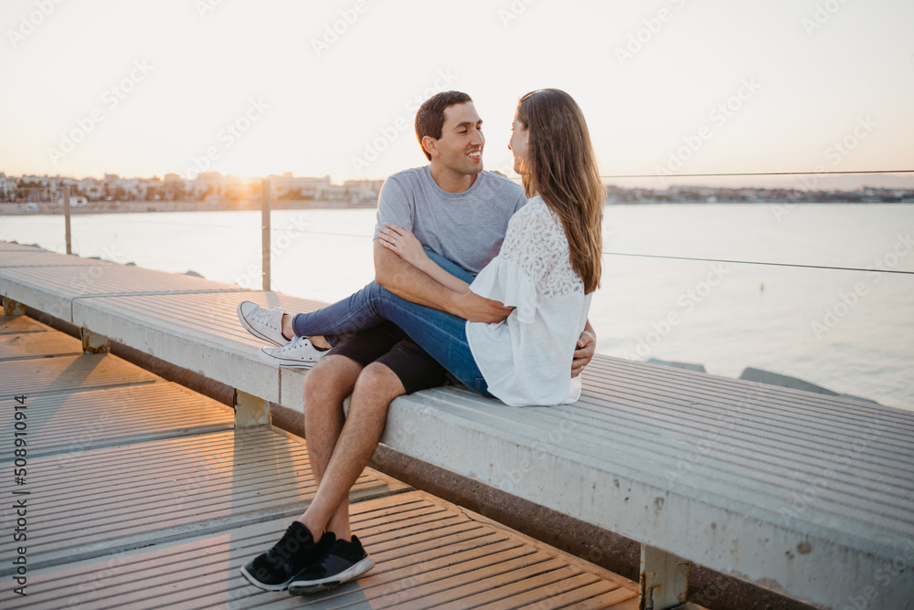 A Hispanic man is hugging his smiling brunette girlfriend on a breakwater in Spain in the evening. A couple of tourists on a date at the sunset in Valencia.