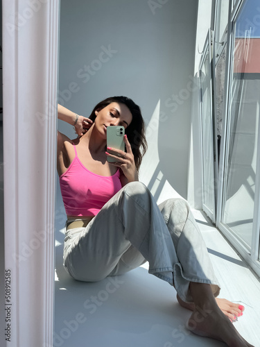 Fit tanned woman in jeans and pink crop top, summer street wear, take photo selfie on phone in mirror for social media, stories, vertical.