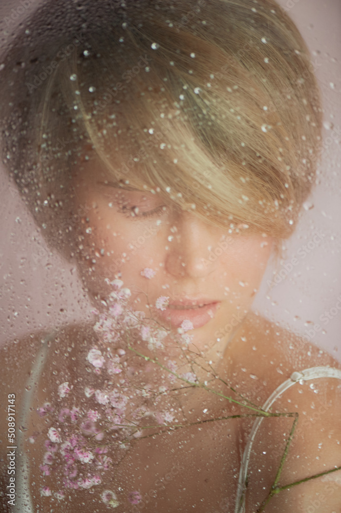 A girl with flowers behind wet glass. Nega and enjoyment of life. Eyes closed. Beauty and personal care industry.