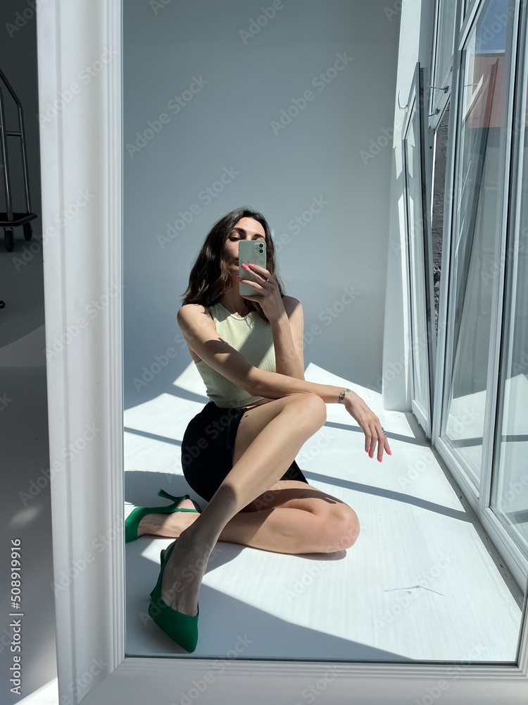 Obraz premium Fit tanned woman in classic short black skirt, light green crop top and heels, summer fashion stylish wear, take photo selfie on phone in mirror for social media, stories, vertical.