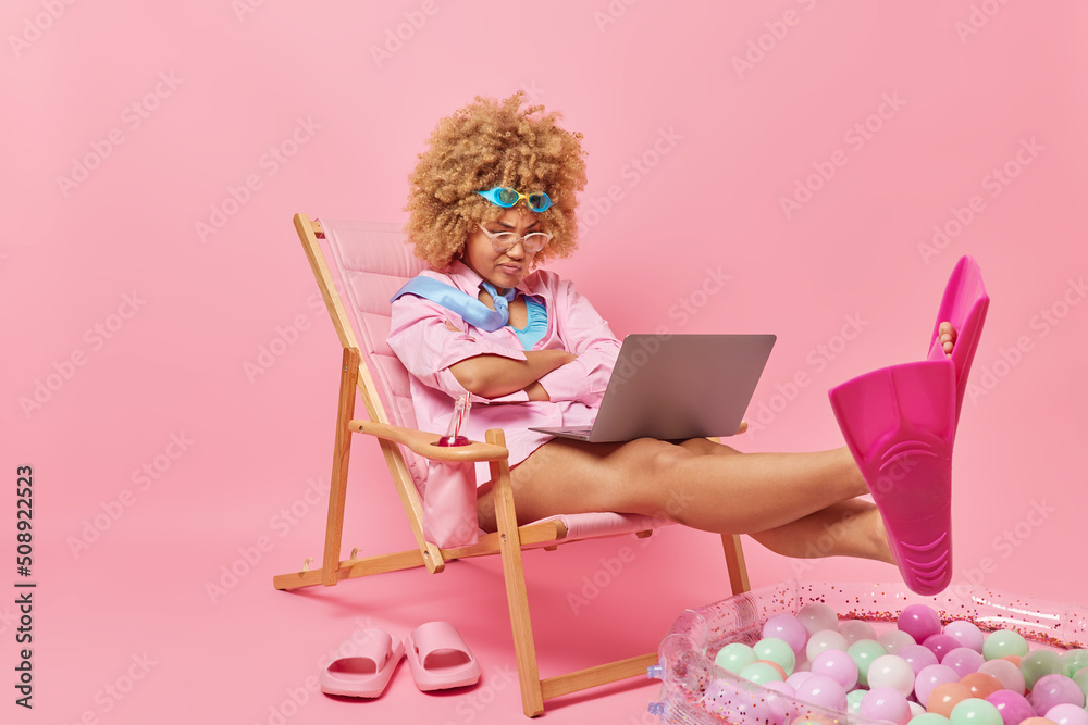 Serious female student focused attentively at laptop screen watches tutorial webinar studies online poses on deck chair being at home but dreams about sea and beach dressed in summer clothes