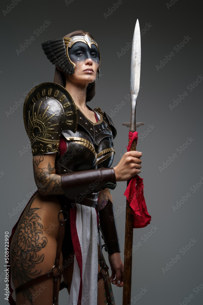 Obraz premium Shot of antique female warrior dressed in steel armor holding spear against grey background.